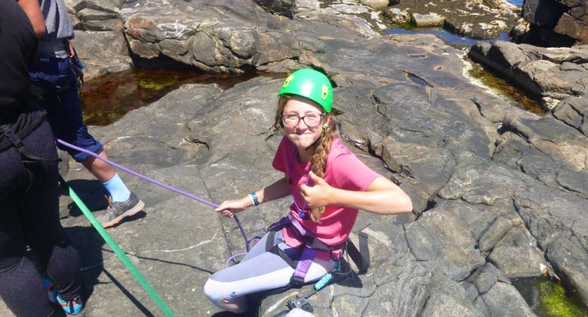A person wearing safety gear sits on a rock and gives the camera a thumbs up